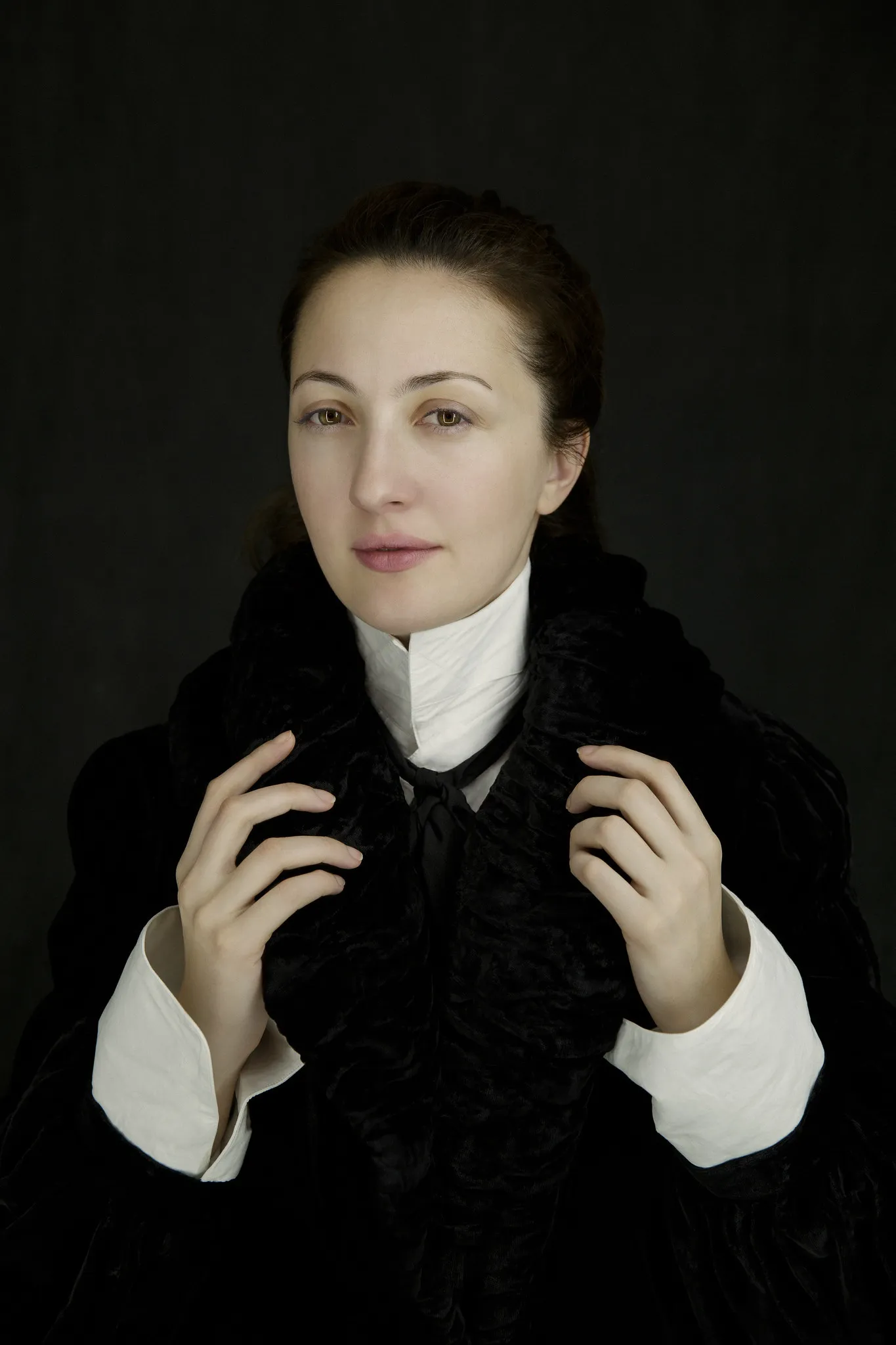 Marika in a black fur collar and white cuffs holds her collar, on a dark background.
