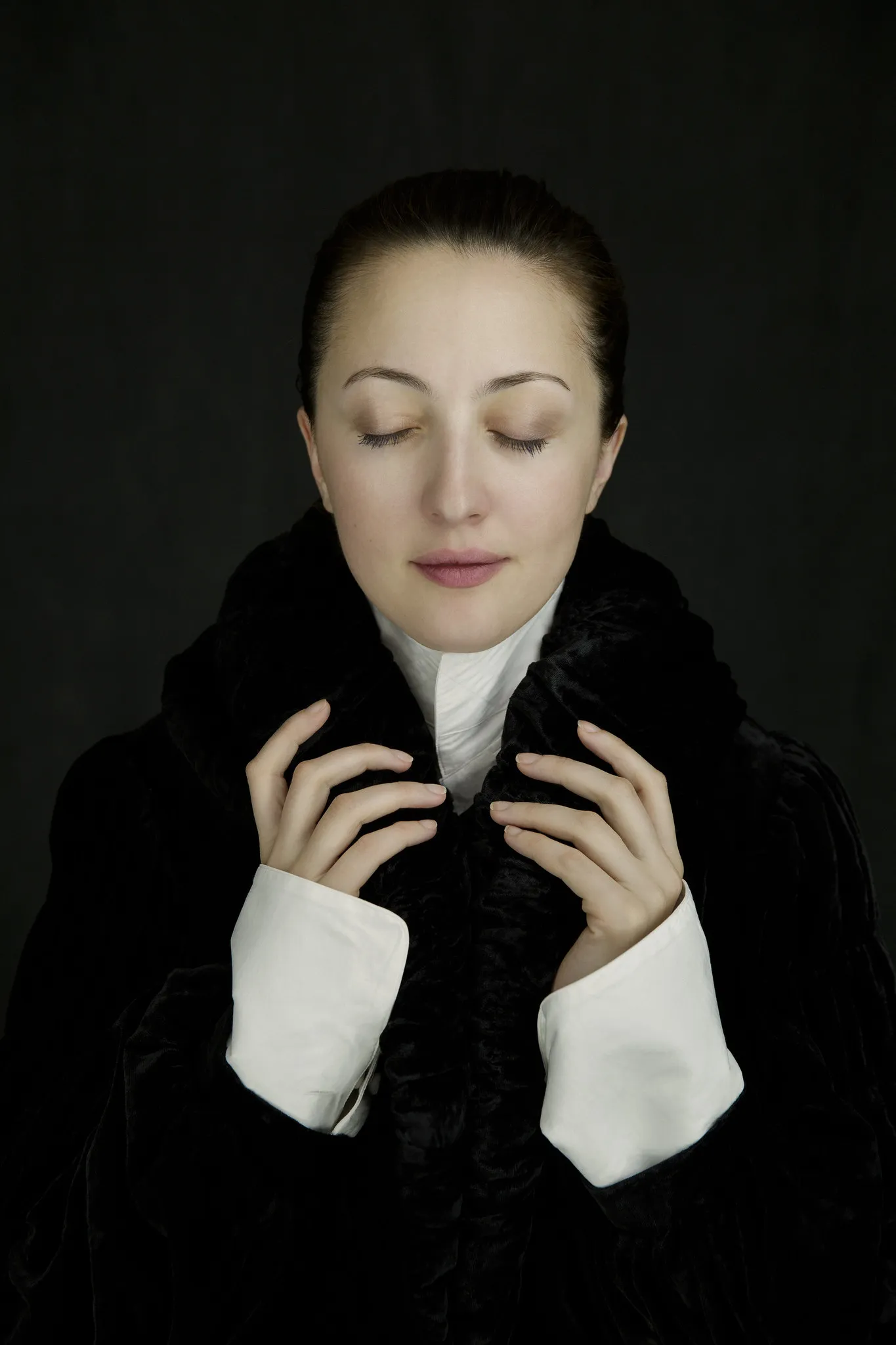 Marika closes her eyes, hands at her fur collar, on a dark background.