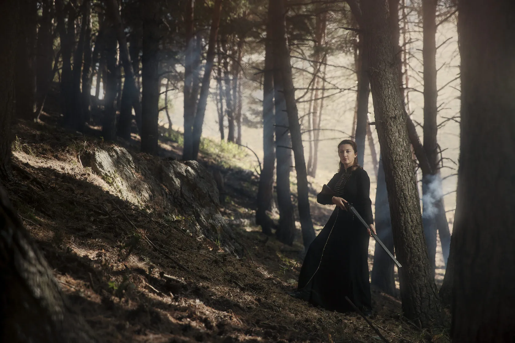 Marika in a long black dress stands on a forest slope, holding a shotgun.