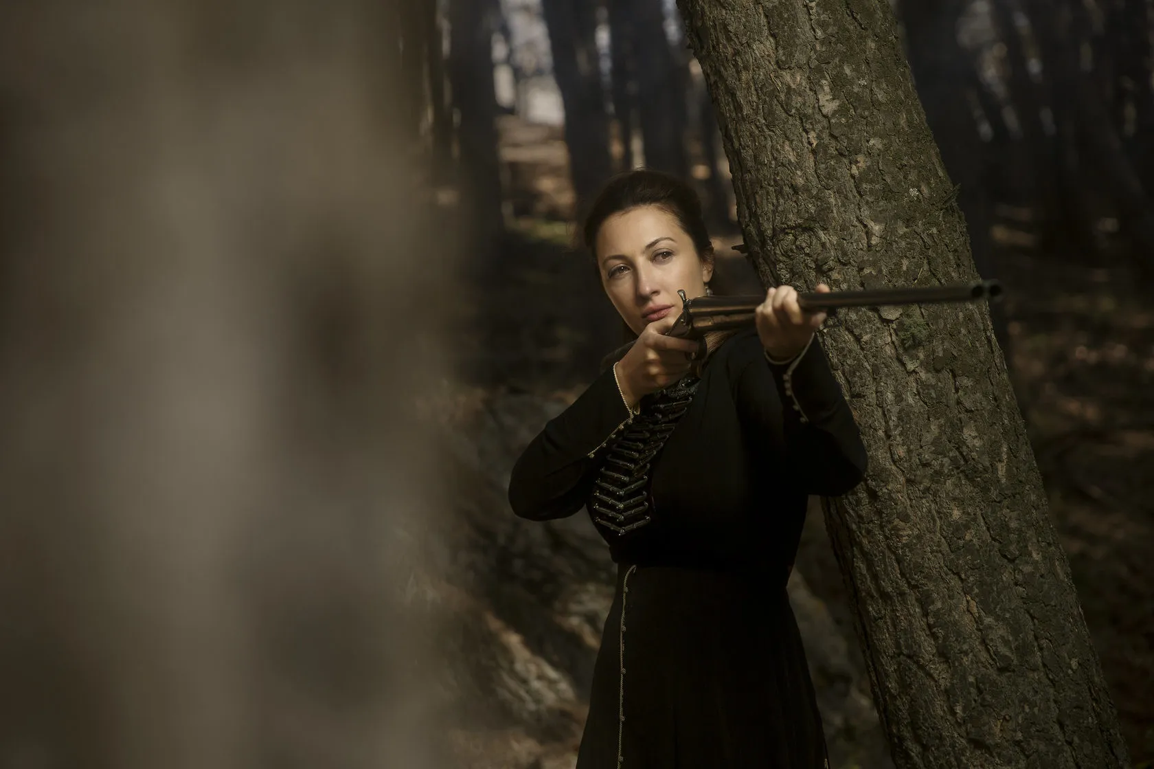 Marika aims a shotgun from behind a pine trunk.
