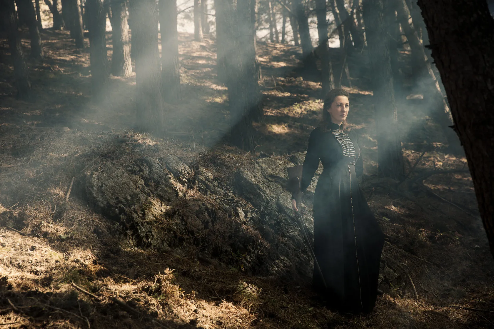 Marika stands in a smoky pine forest, sunbeams falling through the trees.