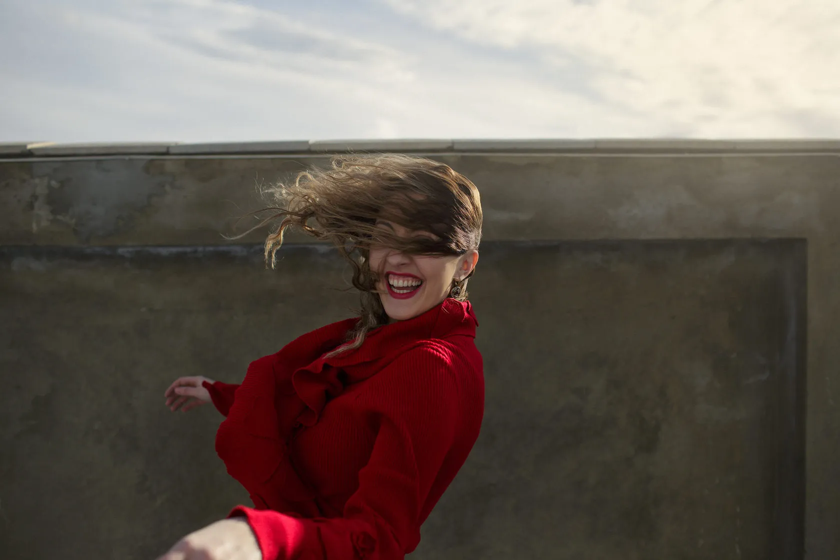 Marika laughs in a red dress by a concrete wall, the wind throwing her hair across her face.