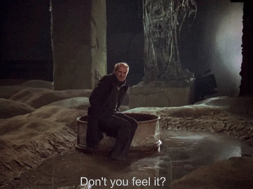 A film still subtitled “Don’t you feel it?”: a man sits on a metal rim in a dark, flooded room.