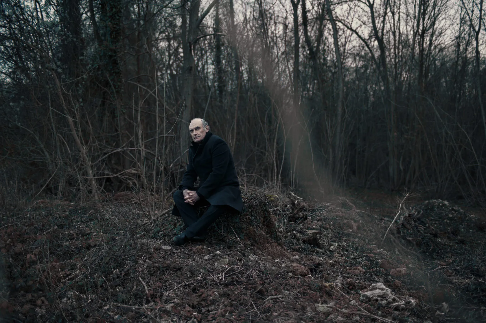 Michel sits on a tree stump in a dark winter wood.