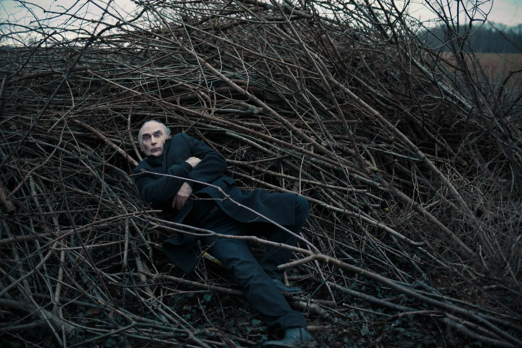 Michel lies stretched out on a pile of branches at the edge of a field.