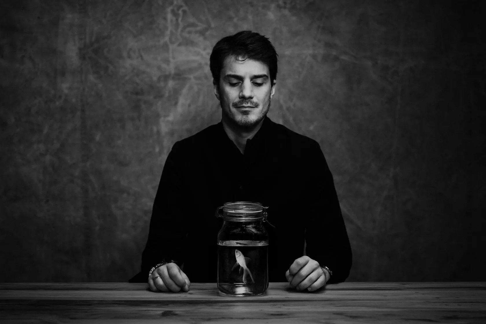 Sven sits at a table, looking down at a fish in a glass jar, in black and white.