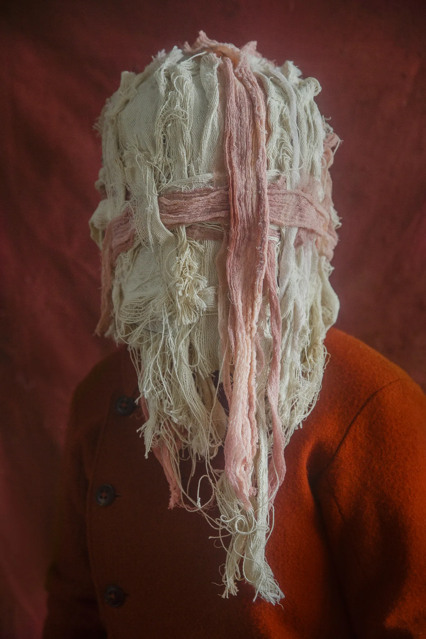 A mask of frayed cream and pink cotton strips covers a head above an orange wool coat.