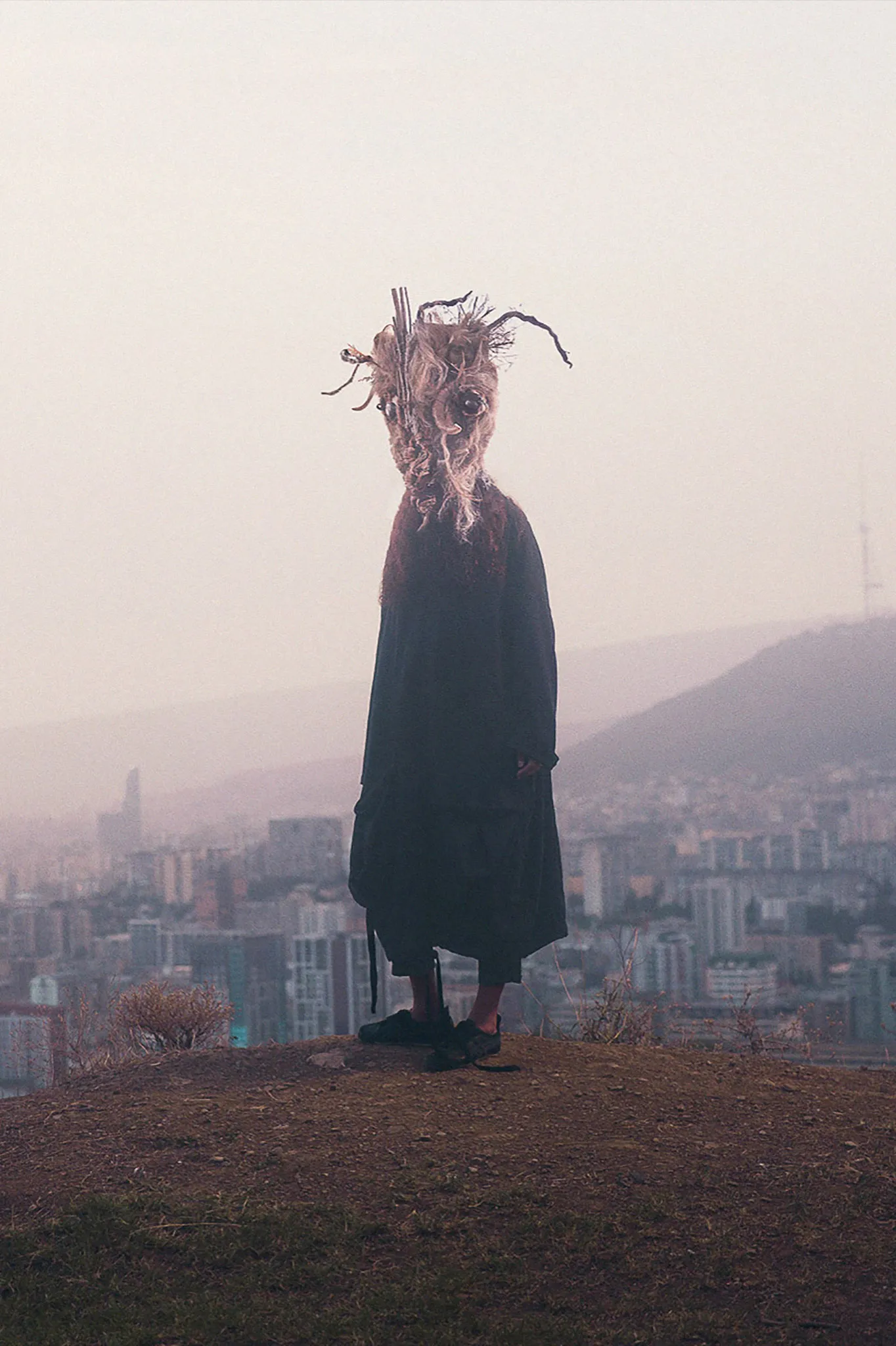 A figure in a long black coat and a shaggy fleece mask stands on a hill above a hazy city.