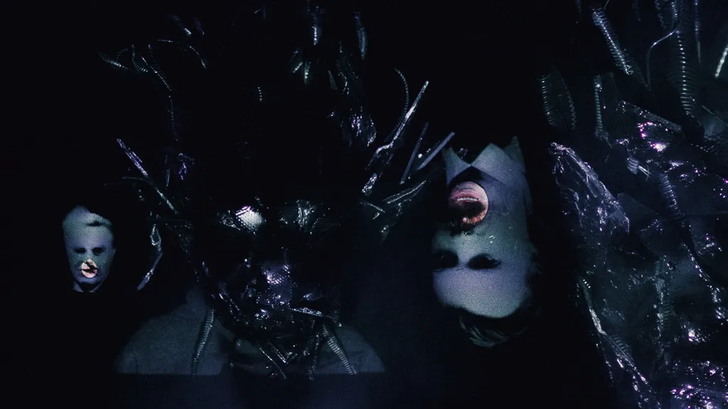 Pale faces, one upside down, float around the dark Chaos mask.