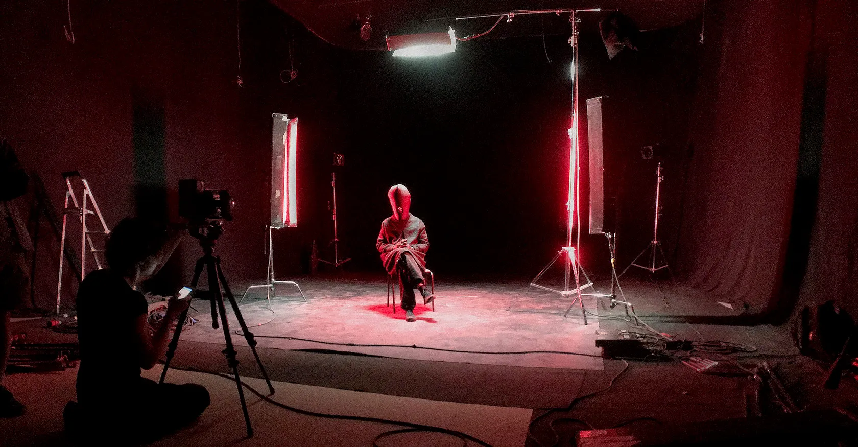 Behind the scenes: a camera operator films a hooded figure seated in a red-lit studio.