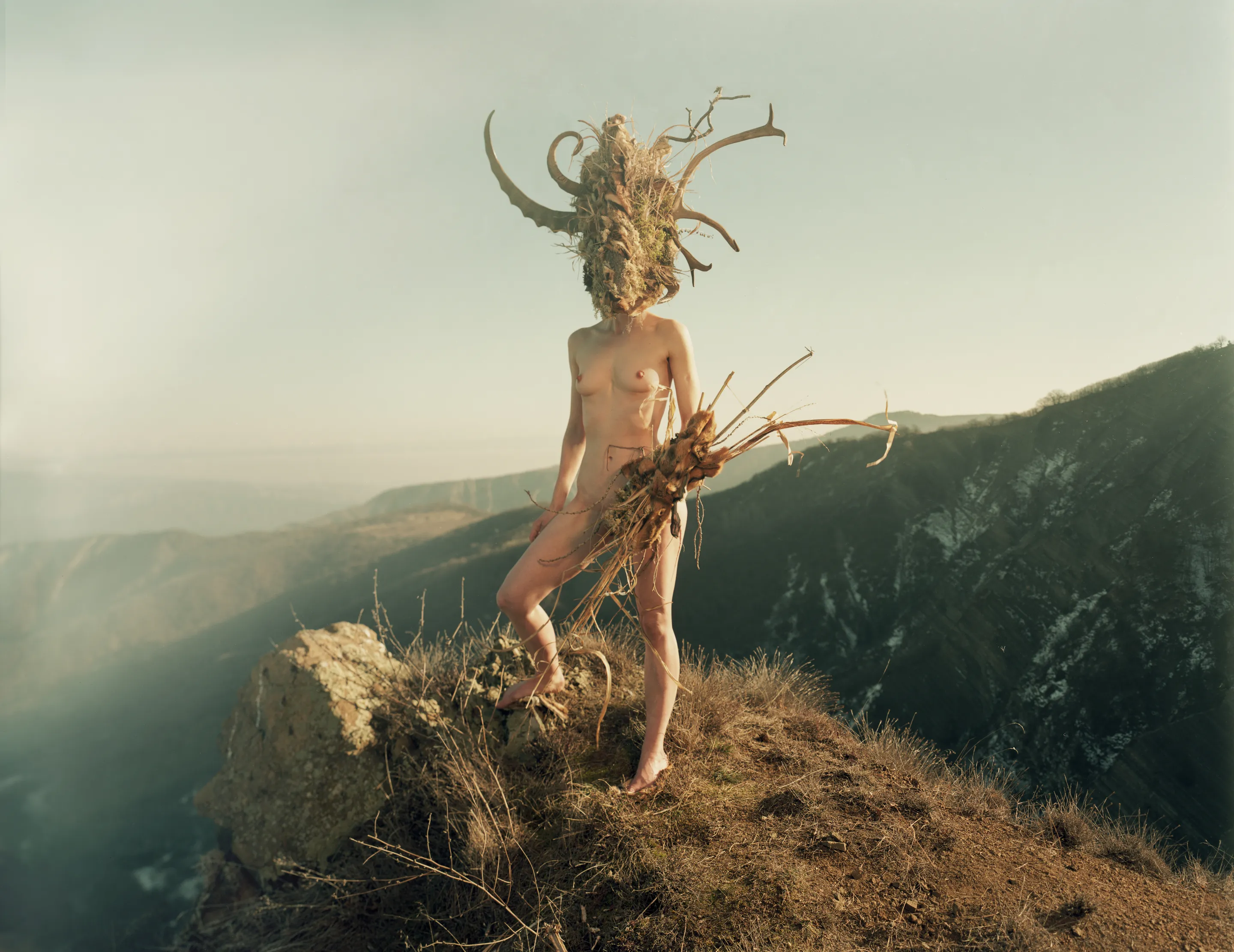 Dàli by Andrei Kovalev: a nude figure crowned with an antler headdress stands on a misty mountain ridge.