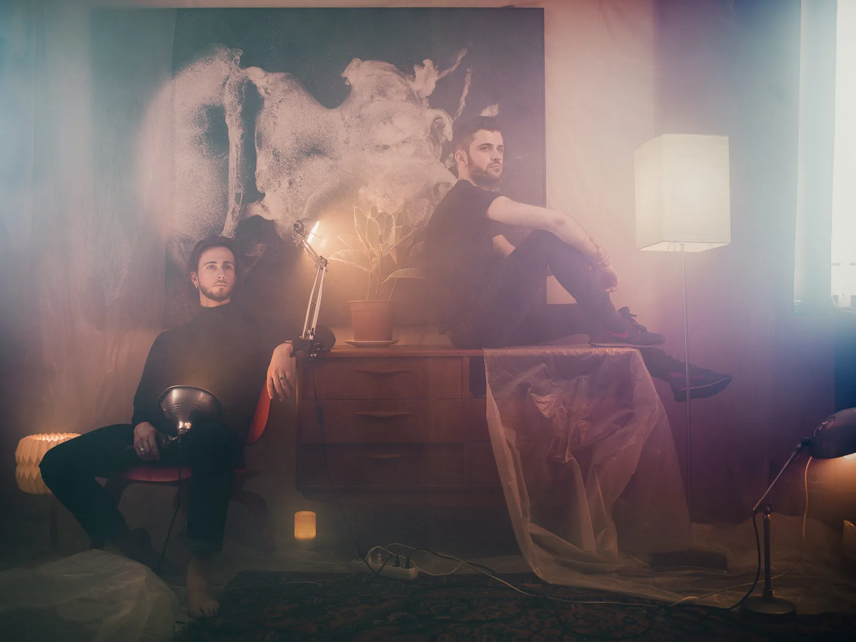 The two members of HYMN by a sideboard among lamps in a hazy room.