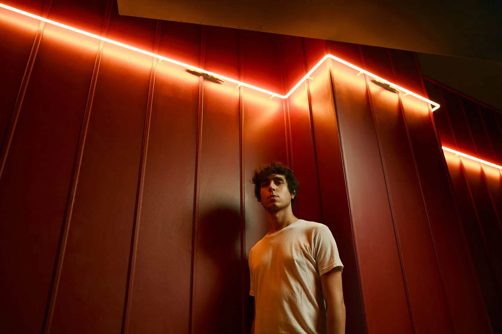 Kazy Lambist stands under a red neon line on a red corrugated wall.