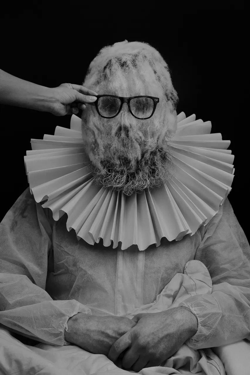 Artiom Krikunov in a paper ruff and a fleece mask, a hand placing glasses on his face.