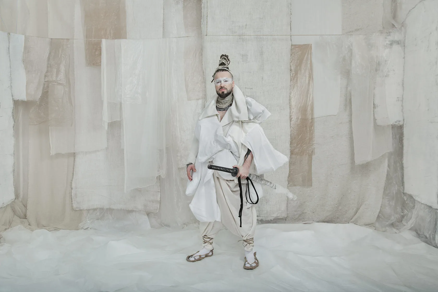 A Burito musician in a white samurai-style costume with a sword before white drapes.