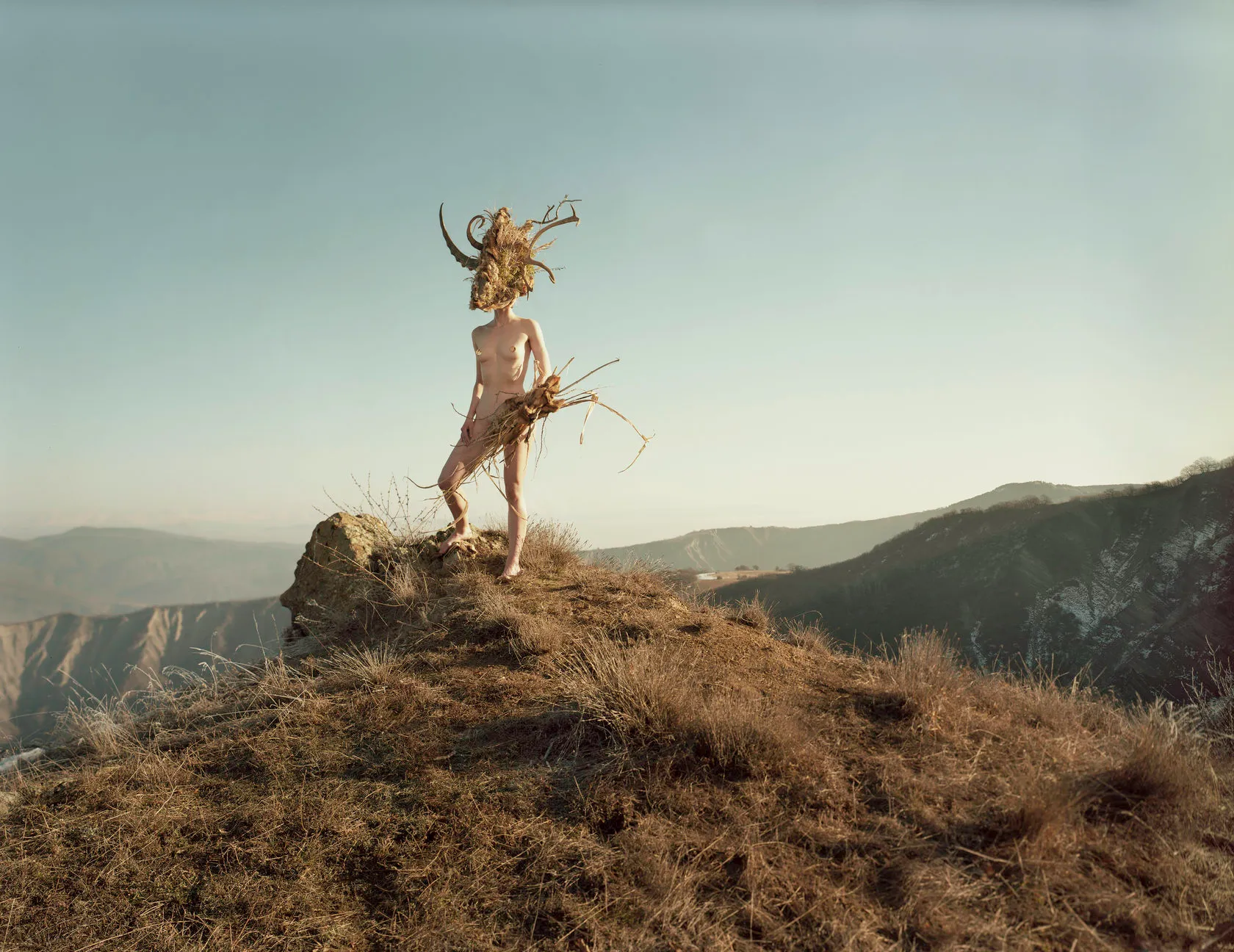 A nude figure in a mask of horns, antlers and dry grass stands on a hilltop holding roots.