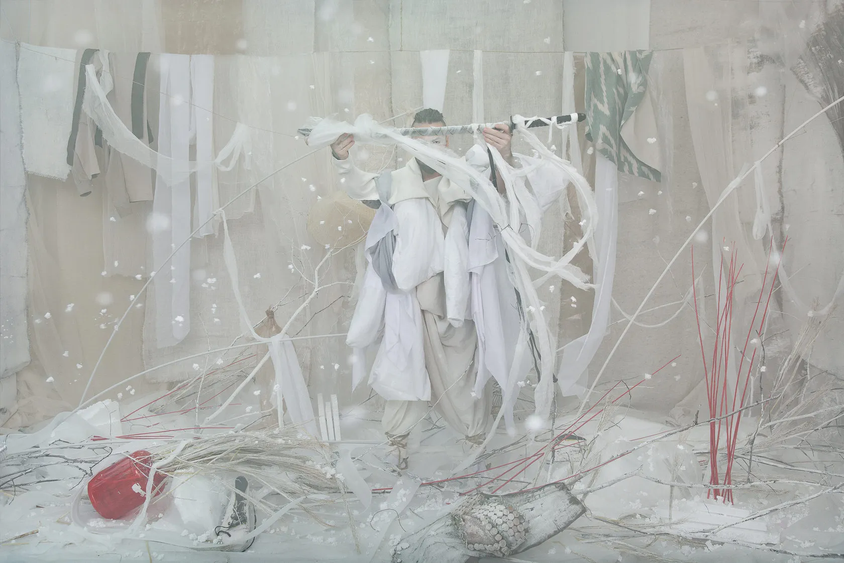 A figure in white carries a pole hung with white ribbons through a snowy set with red sticks.