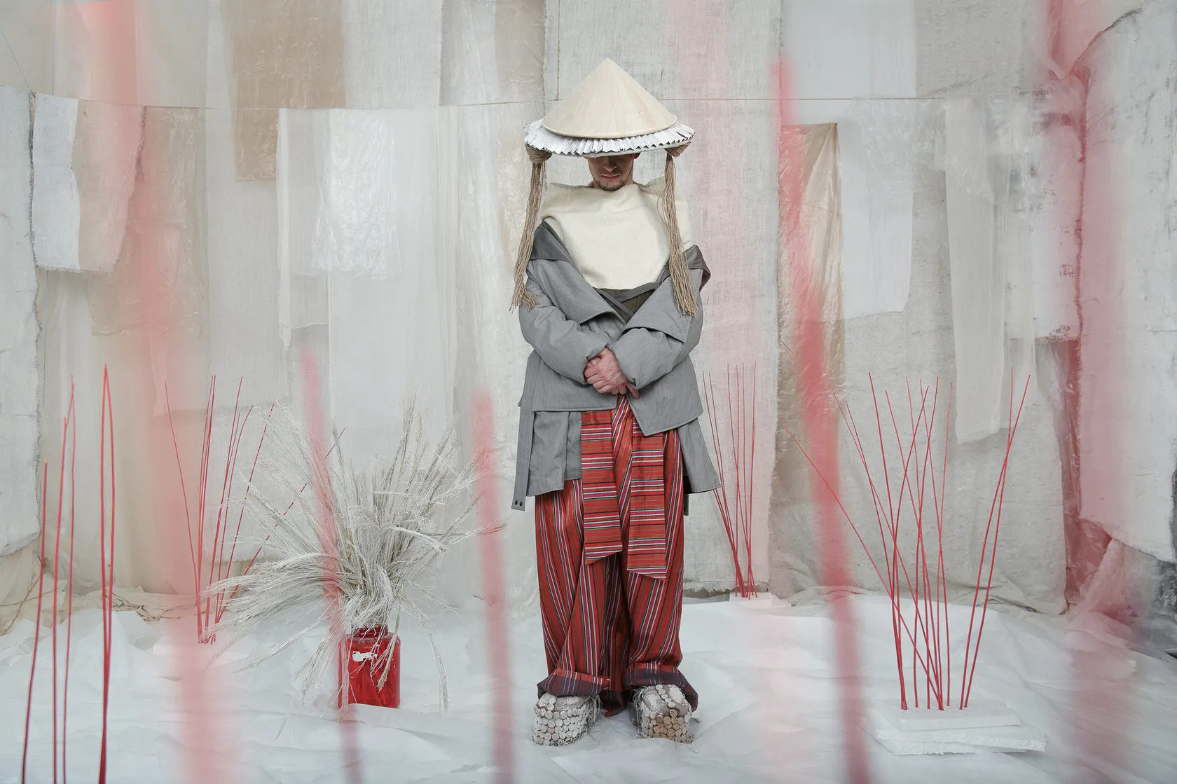 A figure in a veiled straw hat and red striped trousers stands among red sticks.
