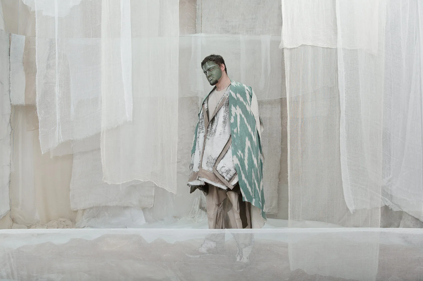 A man with a green-painted face in a teal patterned robe stands behind sheer curtains.