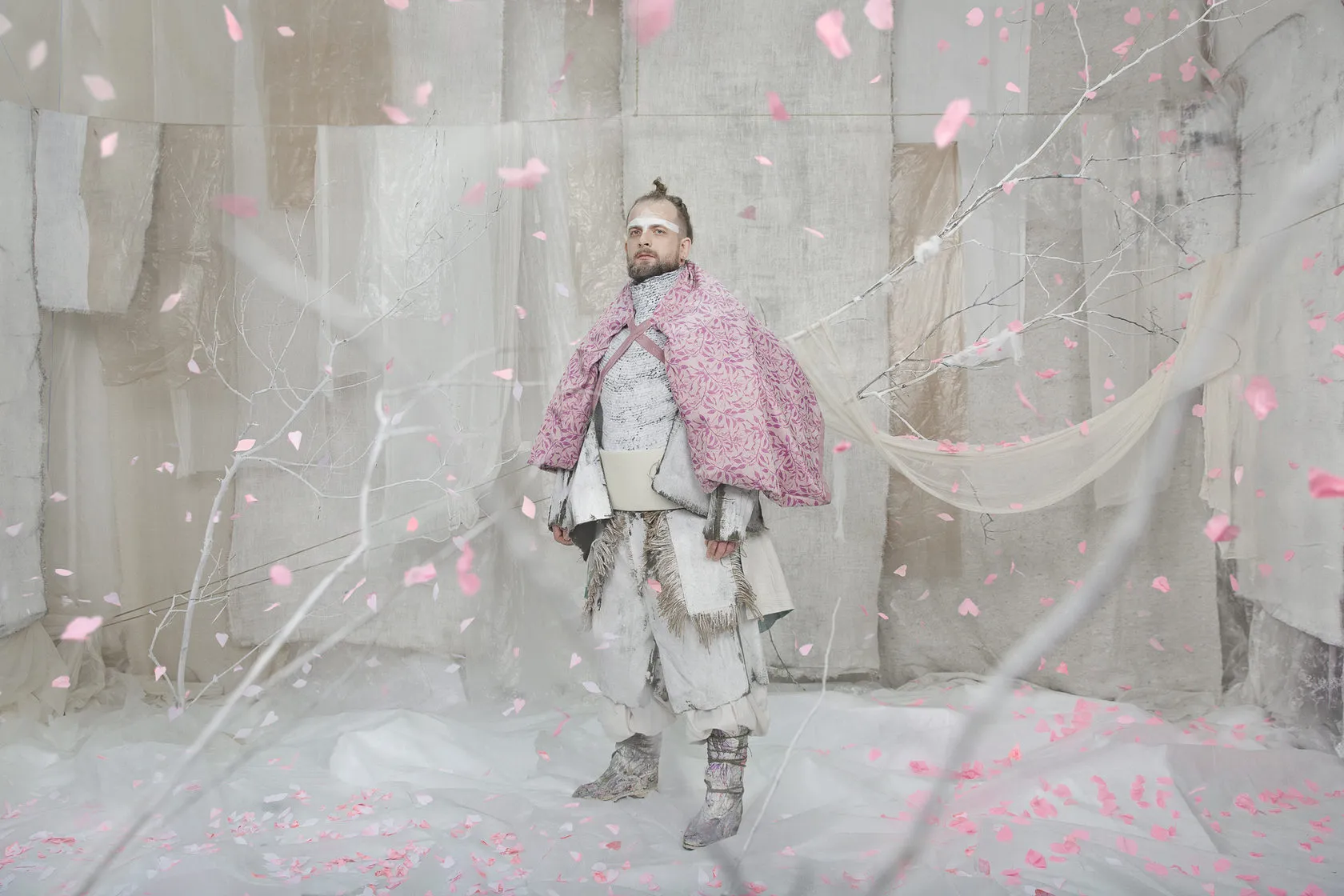 A man in a pink quilted cape stands in a shower of pink petals on a white set.