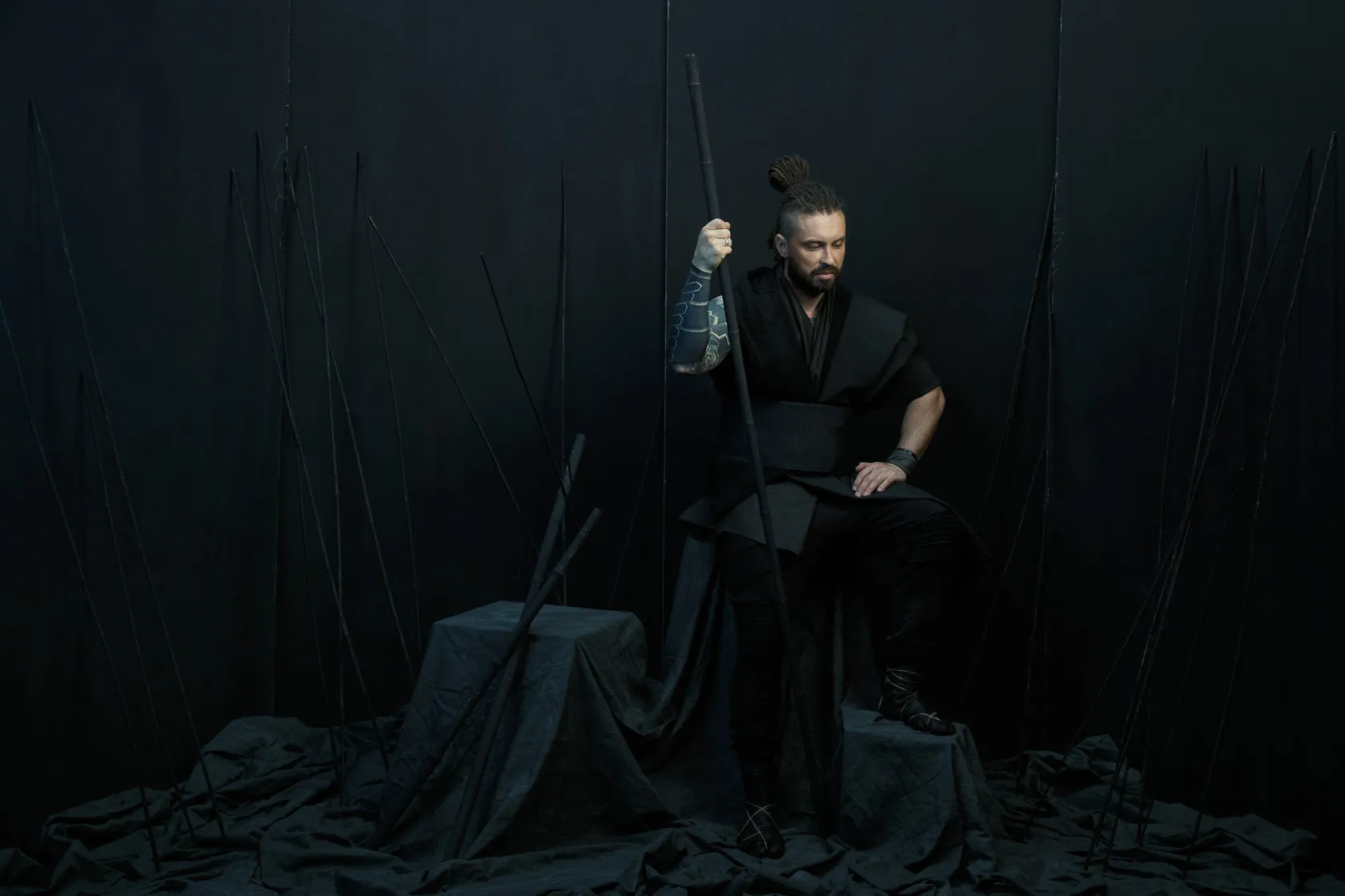 A Burito musician in black sits with a staff on a dark set bristling with poles.