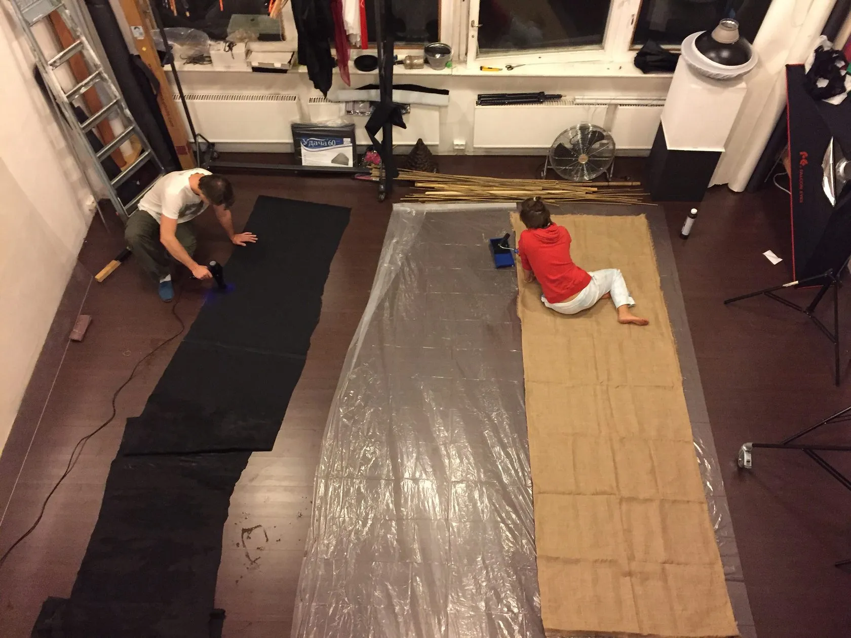Two people work on long strips of black, clear and hessian cloth on a studio floor.