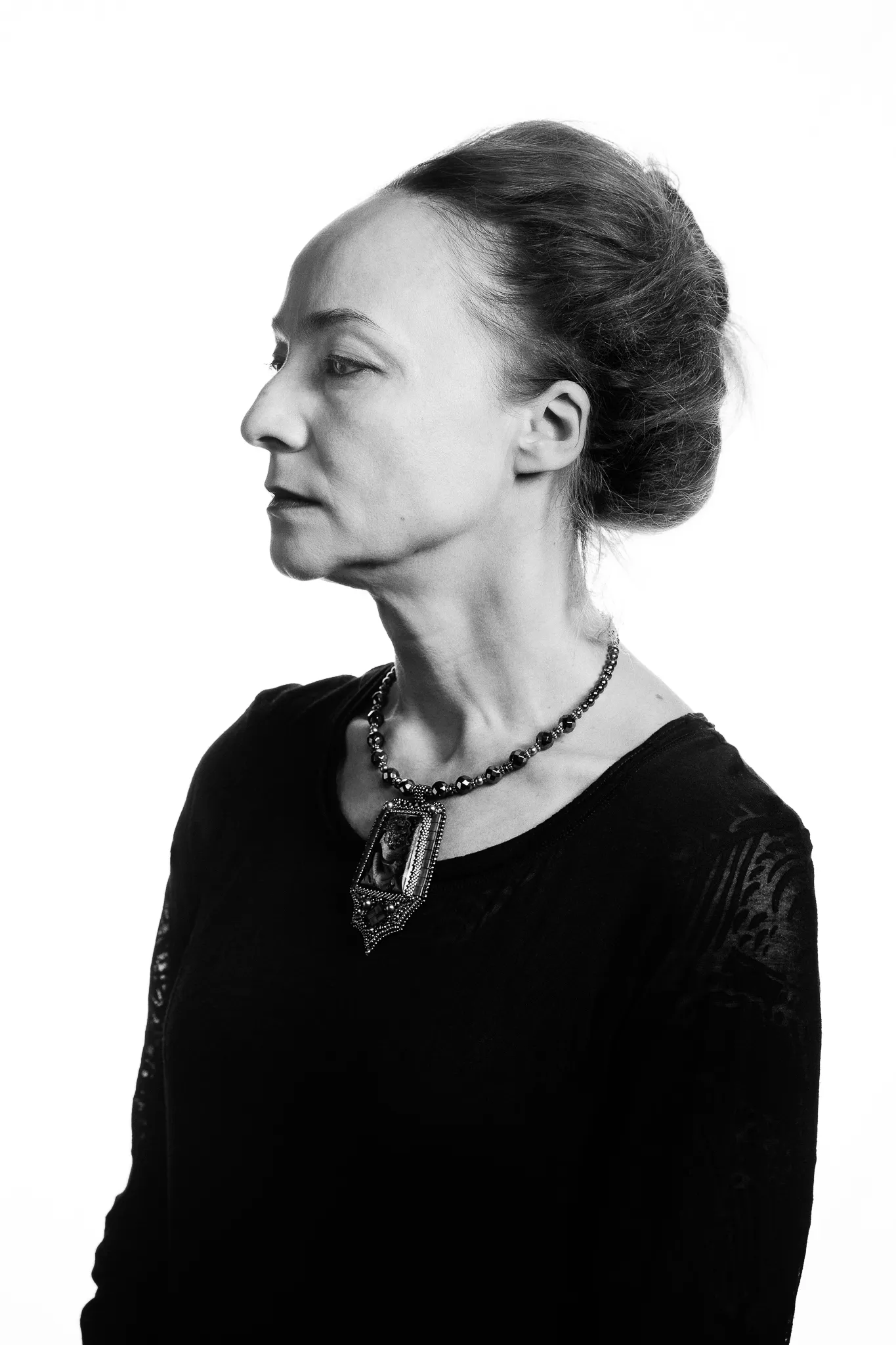 An actress in profile, her hair in a bun, wears a black dress and a long beaded necklace.