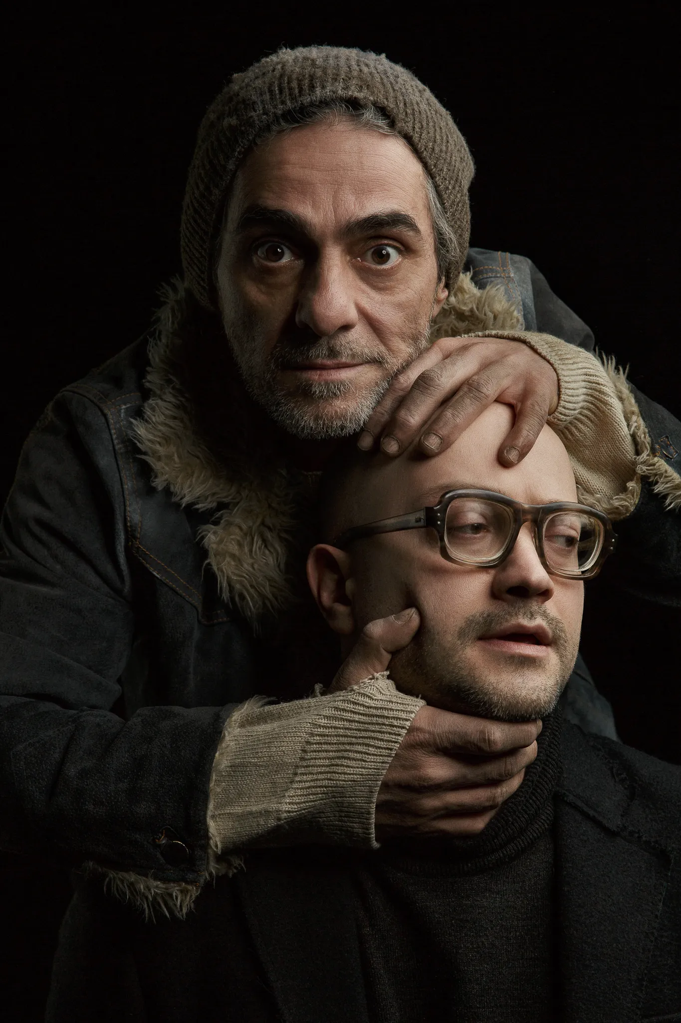 A man in a knit cap holds the head and jaw of a bald man in glasses from behind.