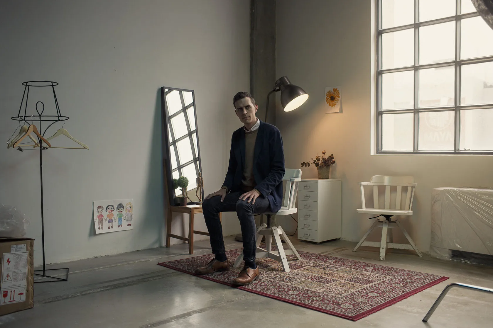 Christian Falsnaes sits on a stool in a sparse studio room with a rug, a mirror and a lamp.