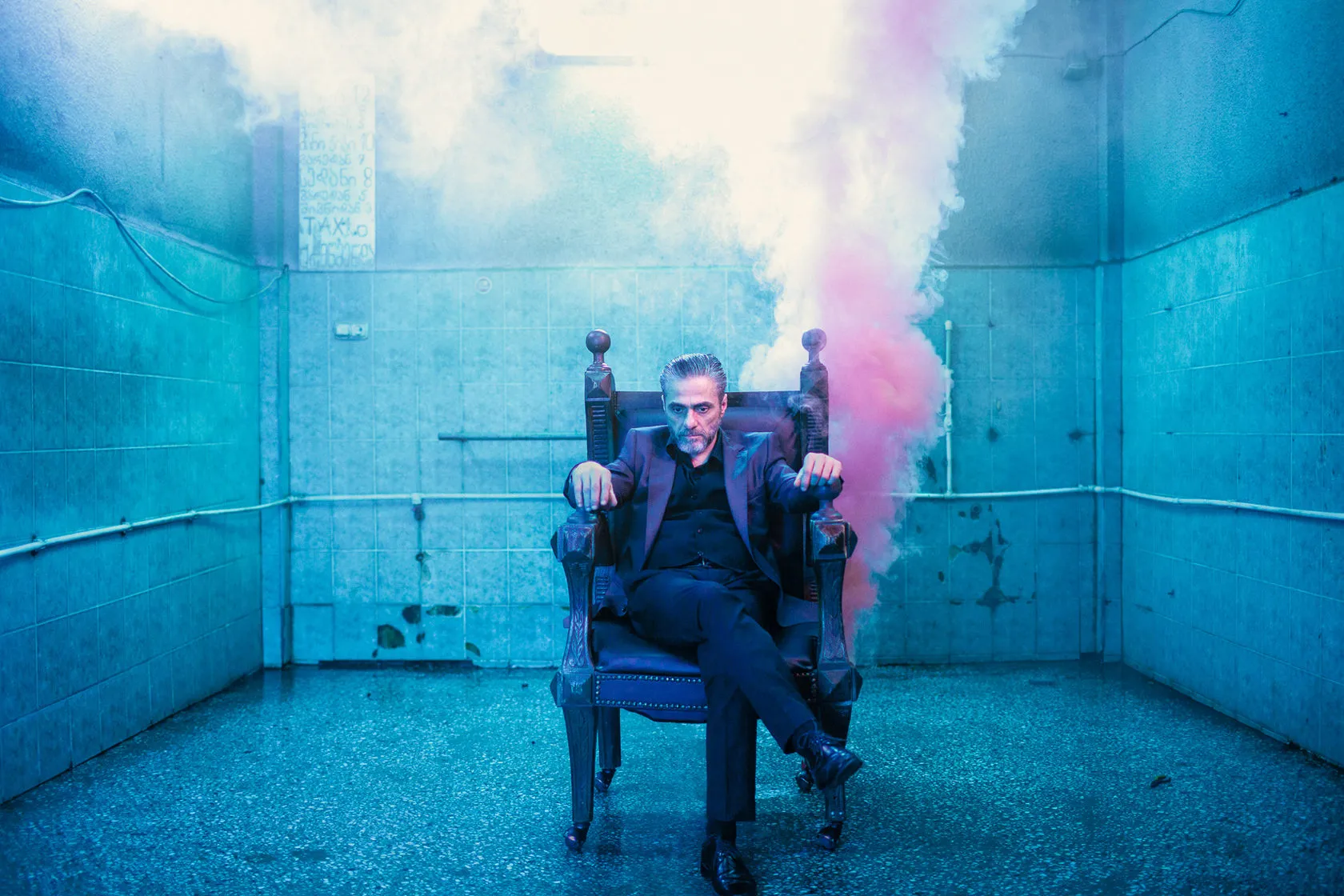 Malkhaz Abuladze as Claudius sits on a throne in a blue tiled room as pink smoke rises behind him.