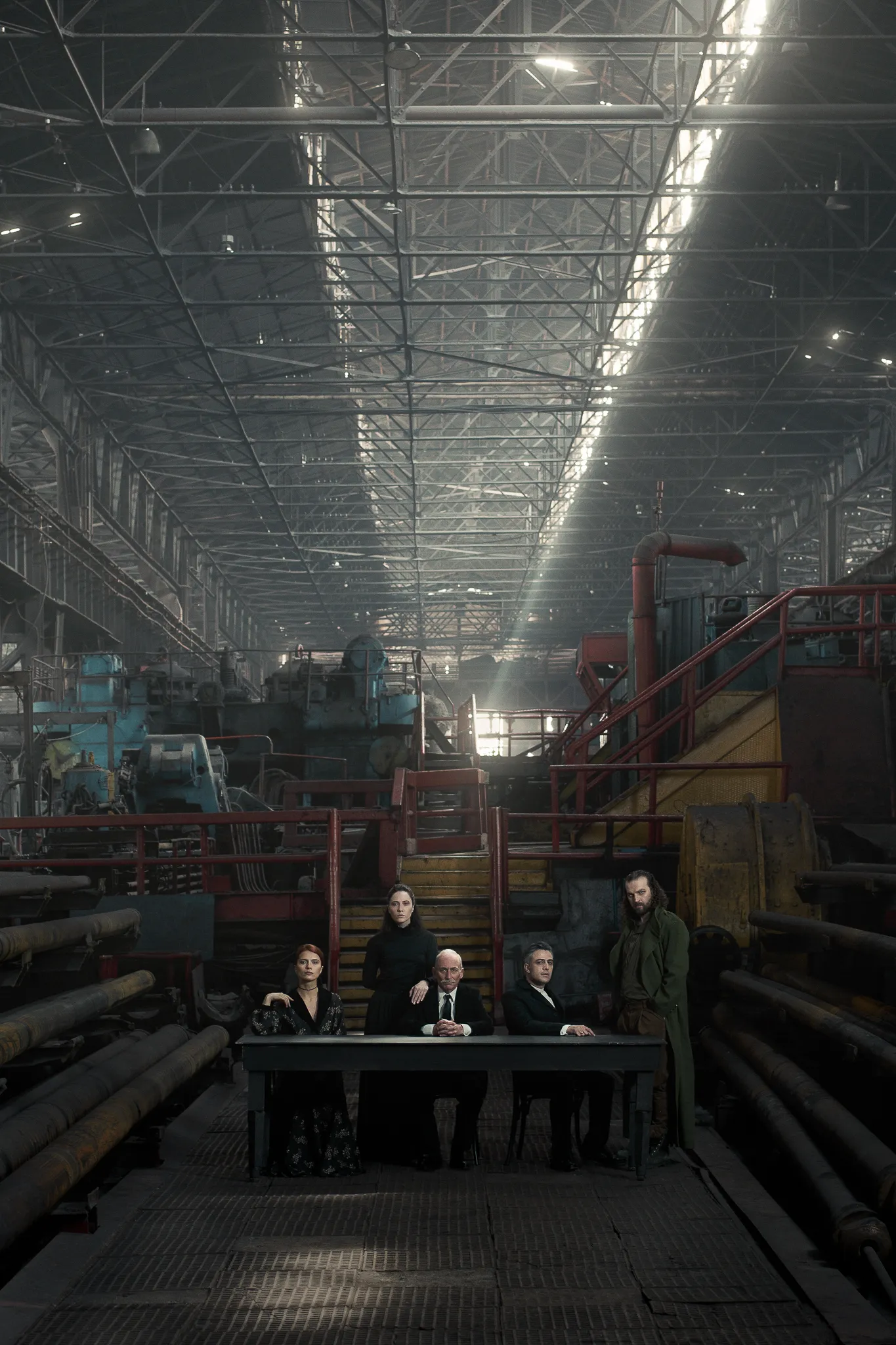 The cast sits at a long table at the far end of a vast factory hall.