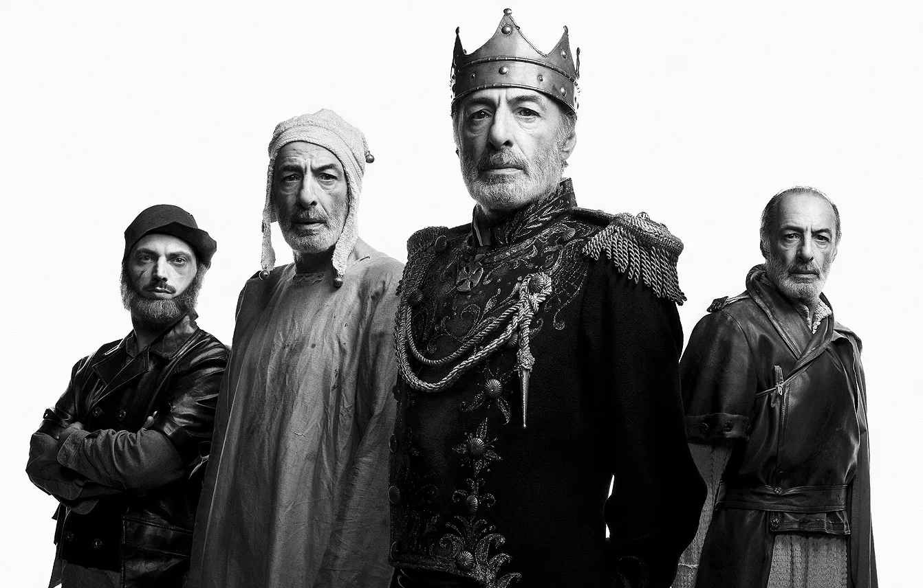 Four actors in costume in black and white, the King in a crown and a braided uniform.