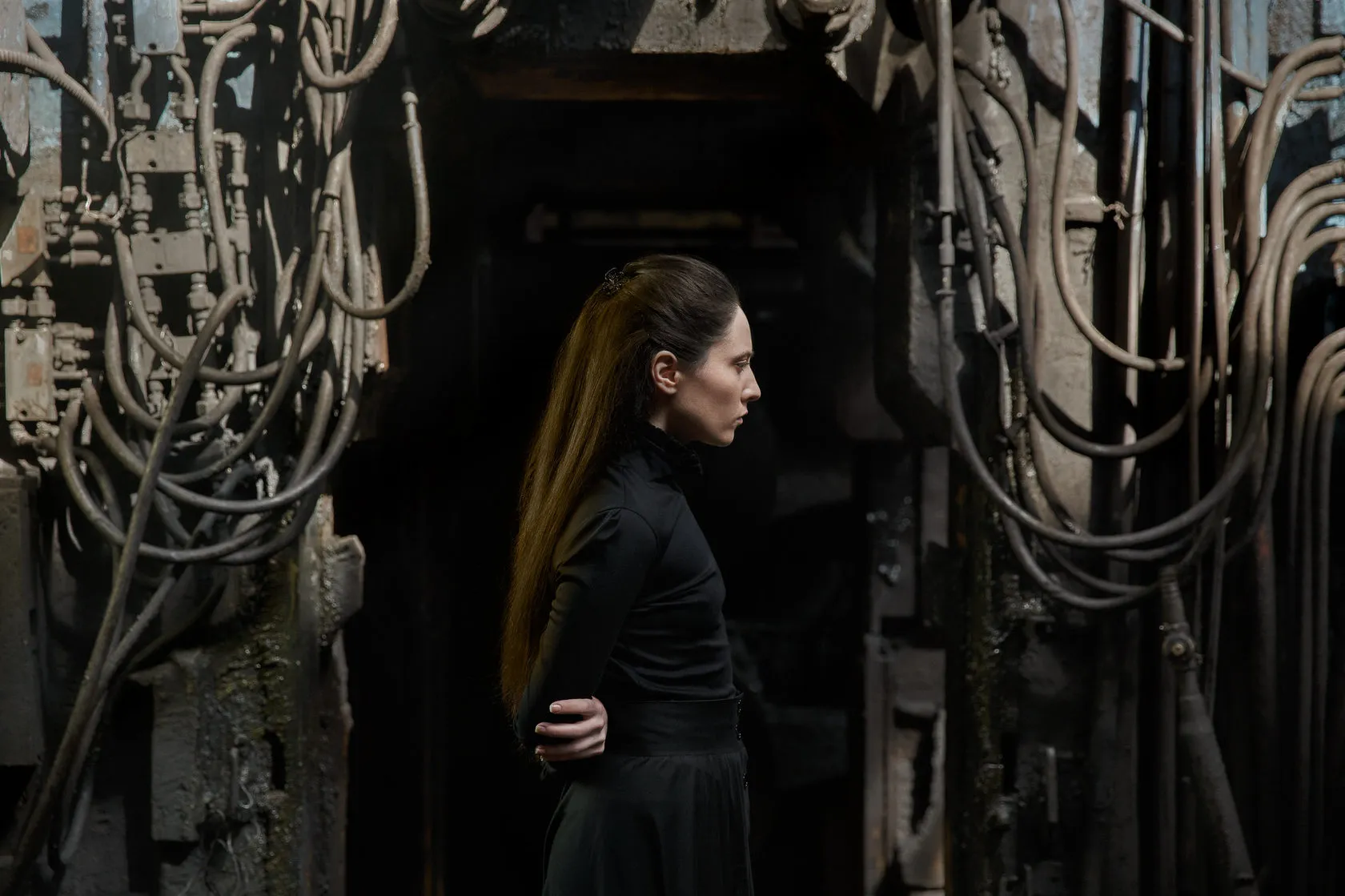 An actress with a long braid stands in profile among tangled cables.