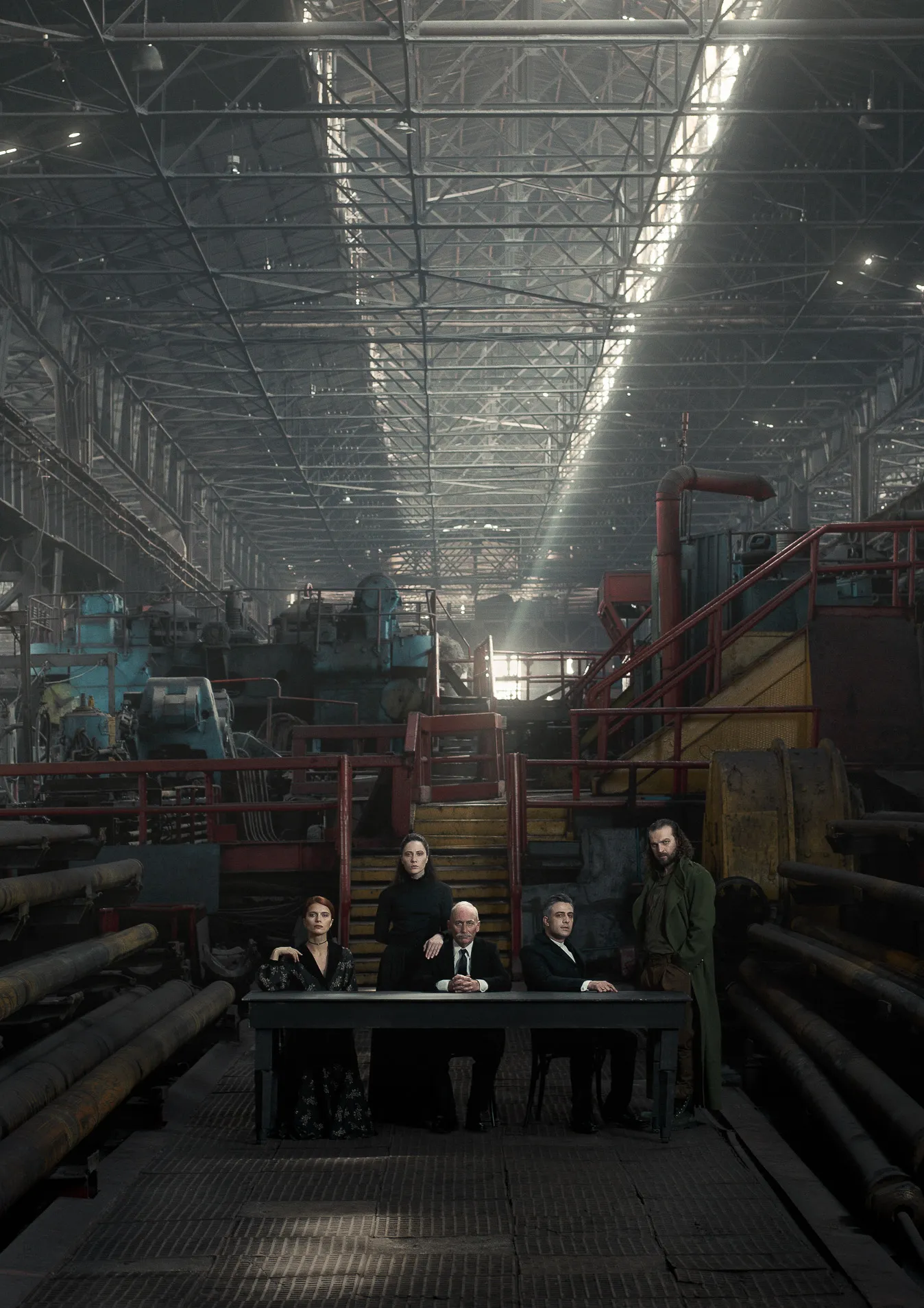 The cast of five sits at a long table in a vast factory hall.