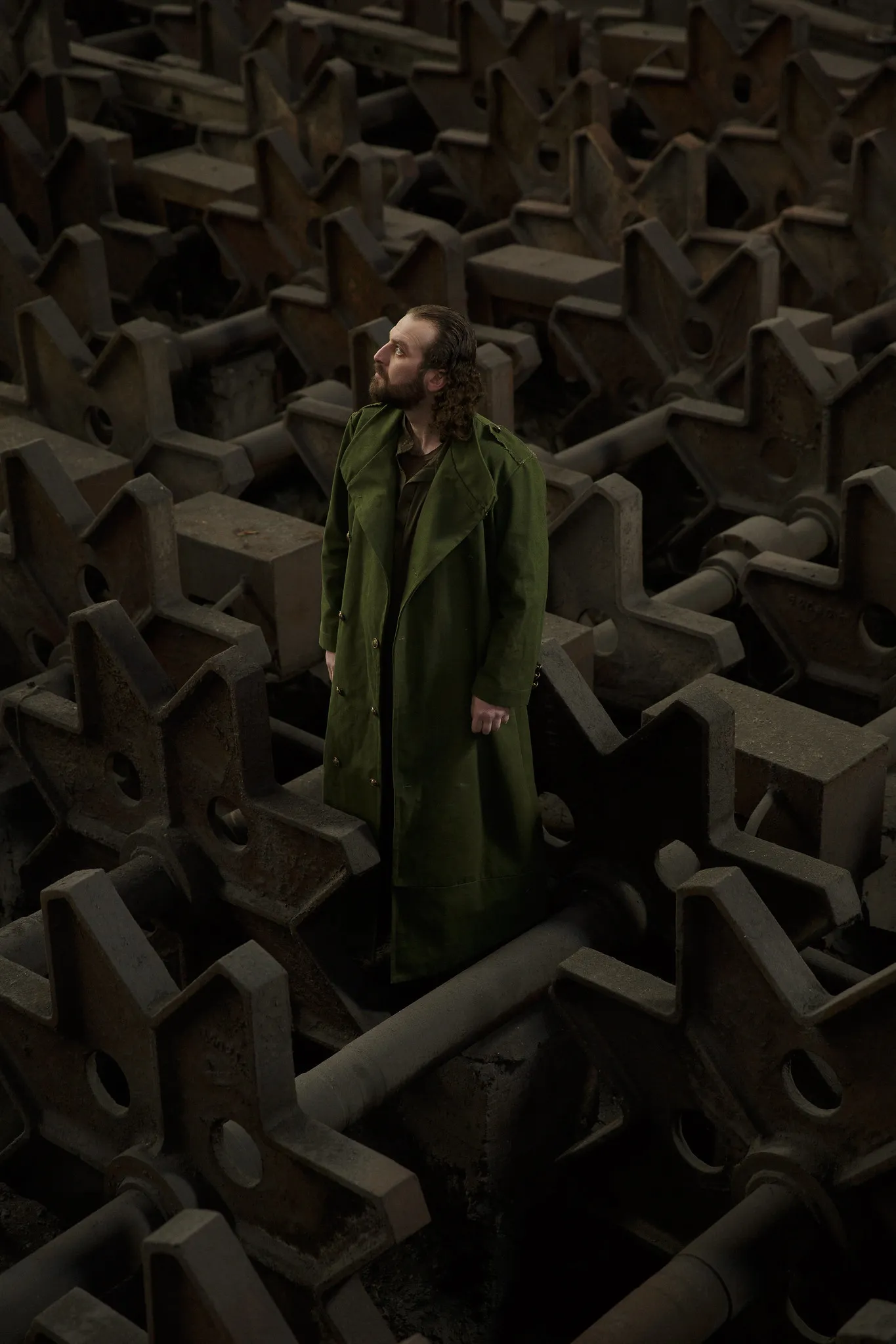 A bearded man in a long green coat stands among heaps of iron cogs.