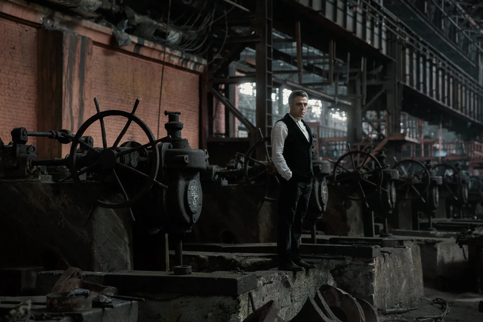 A man in a waistcoat stands by a large iron wheel in the factory hall.