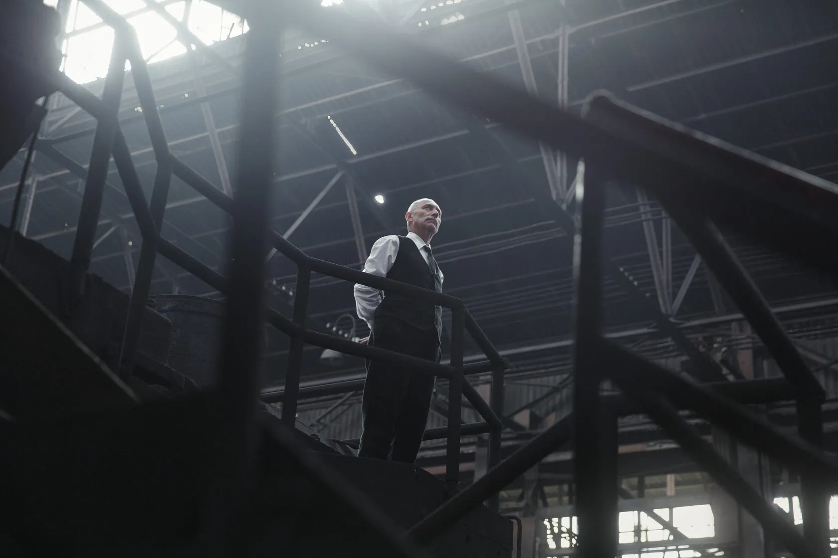 An older man in a waistcoat on a steel staircase, backlit by the factory skylights.