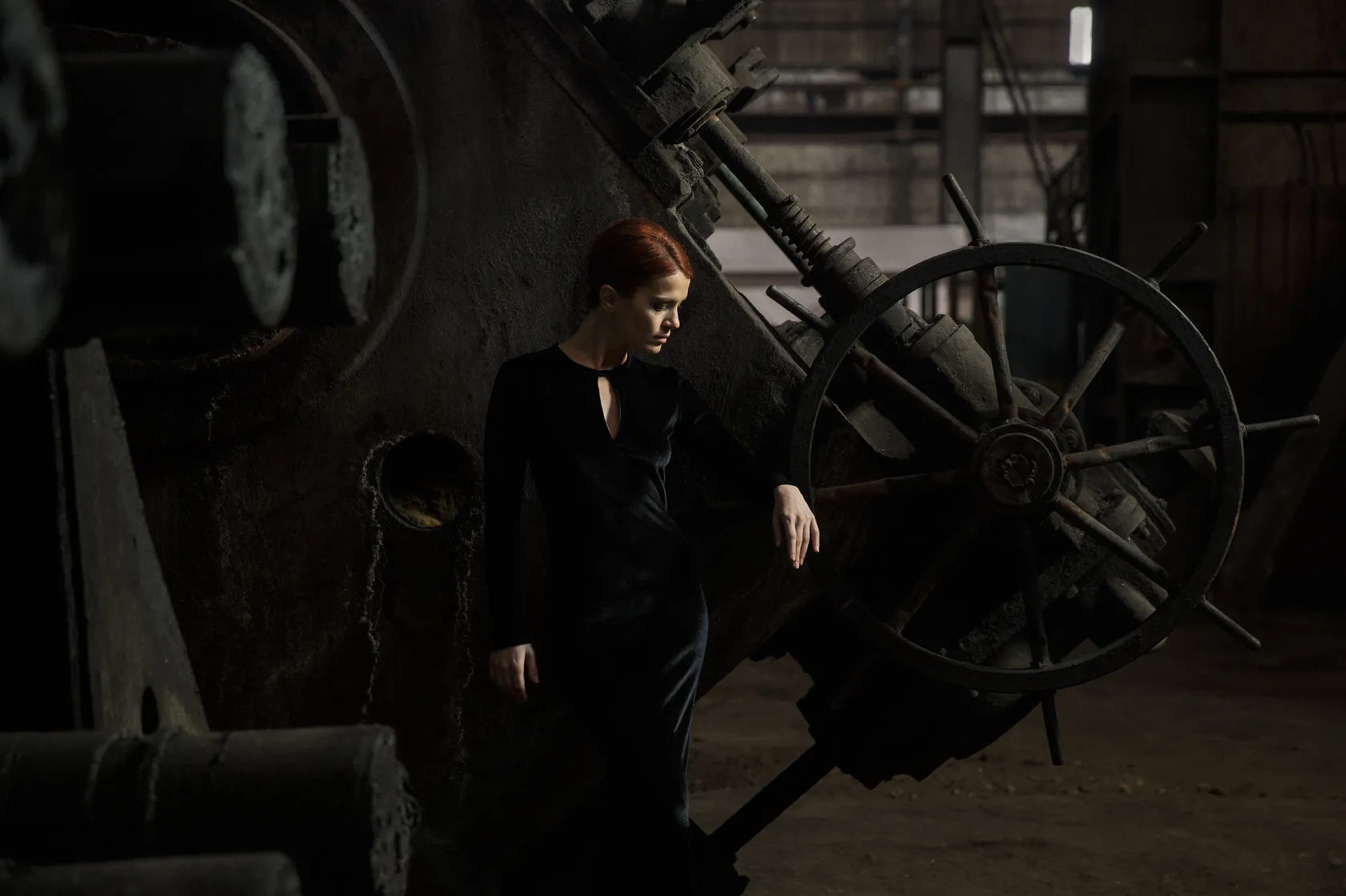 A red-haired actress leans on a huge iron wheel in the dark.