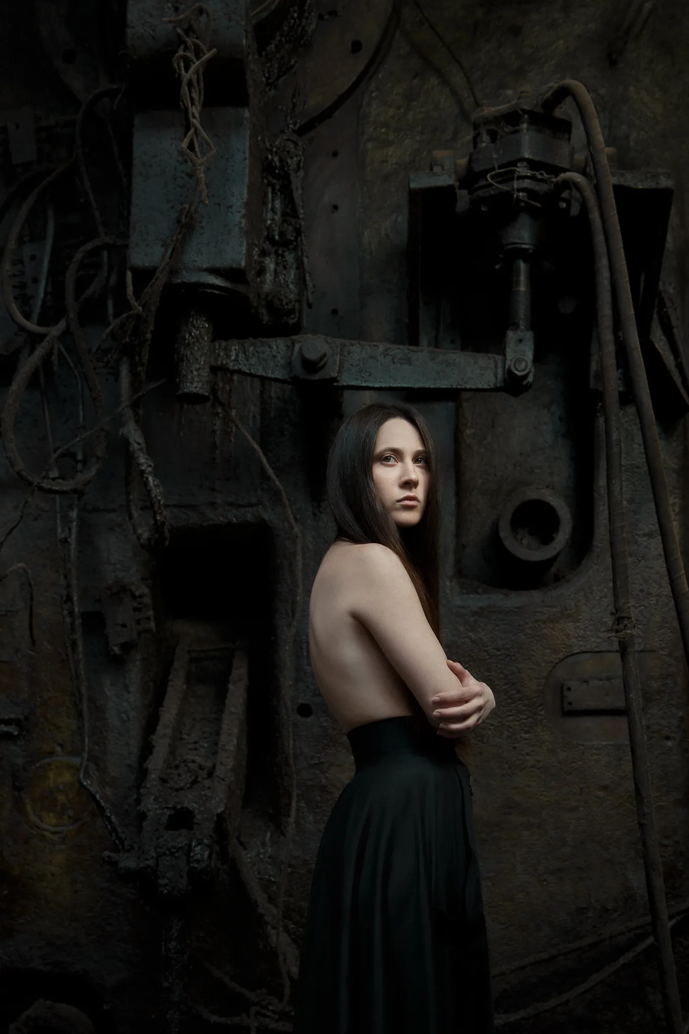 An actress with a bare back stands against a dark iron machine.