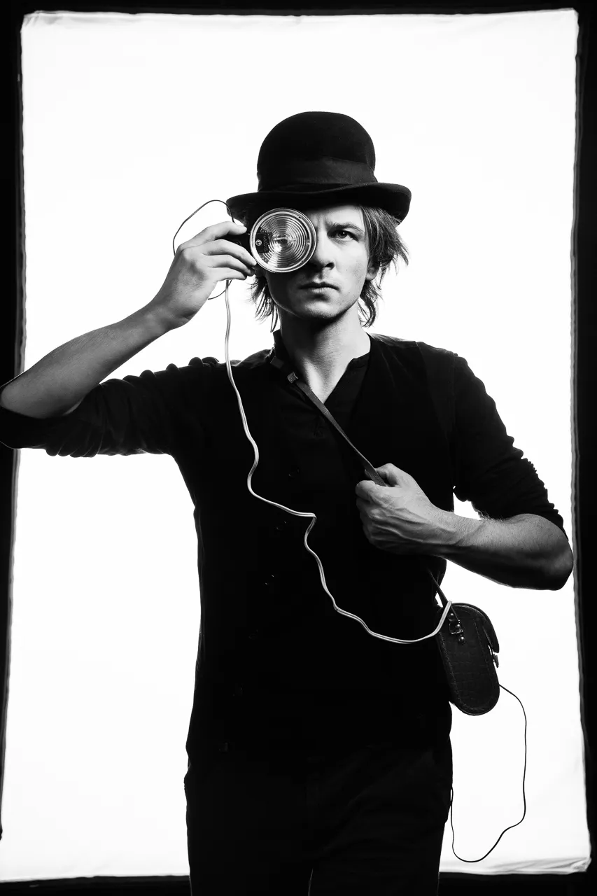 An actor in a bowler hat holds a corded lamp to one eye like a monocle.