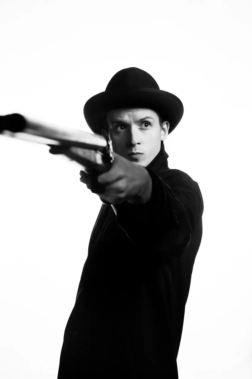 An actor in a bowler hat aims a rifle past the camera.