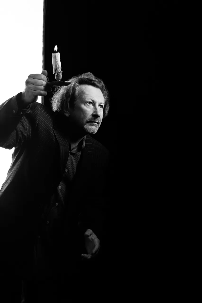 An actor holds a lit candle above his head and peers out of the dark.