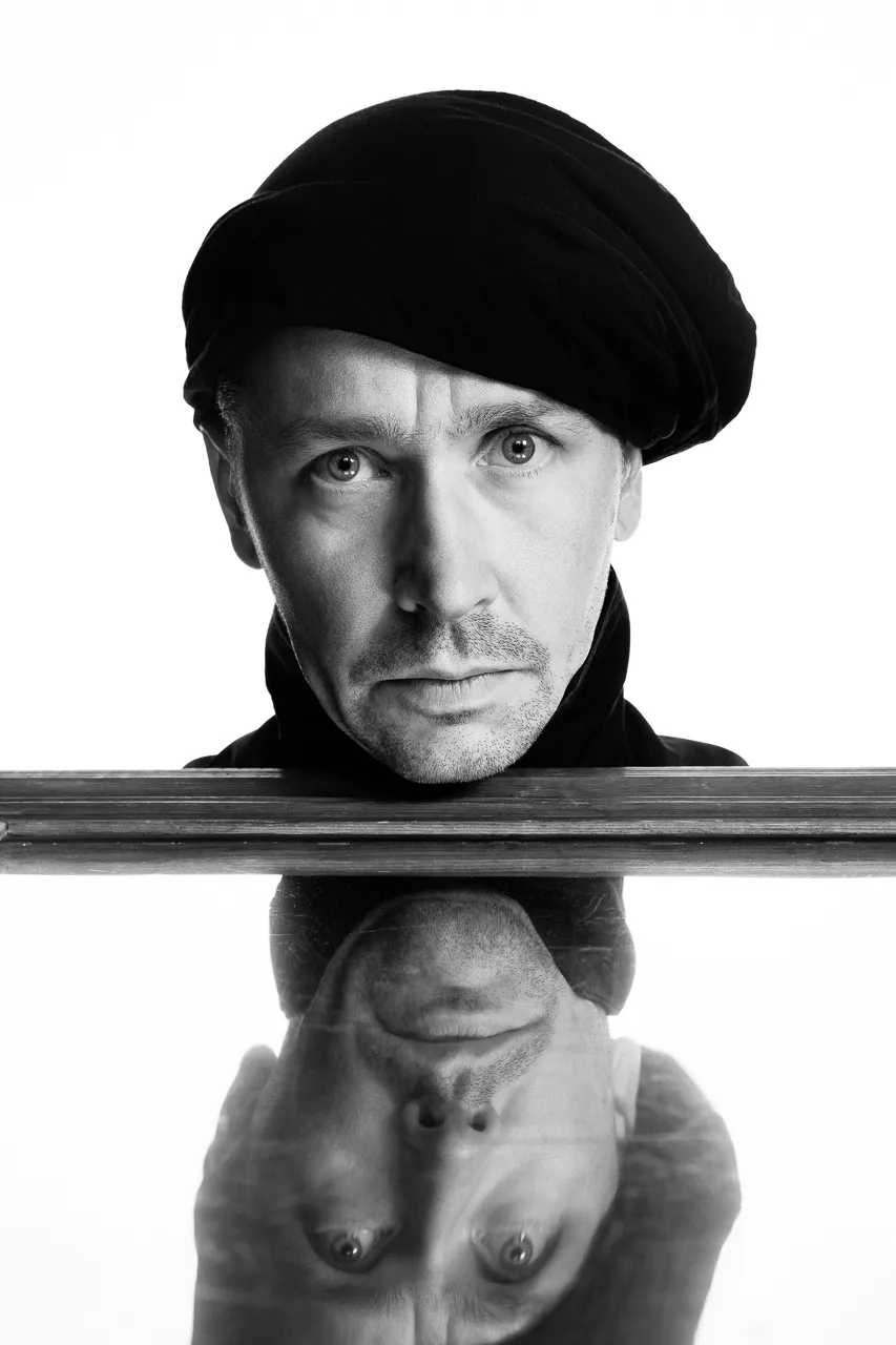 An actor in a black turban rests his chin on a mirror that reflects his face.