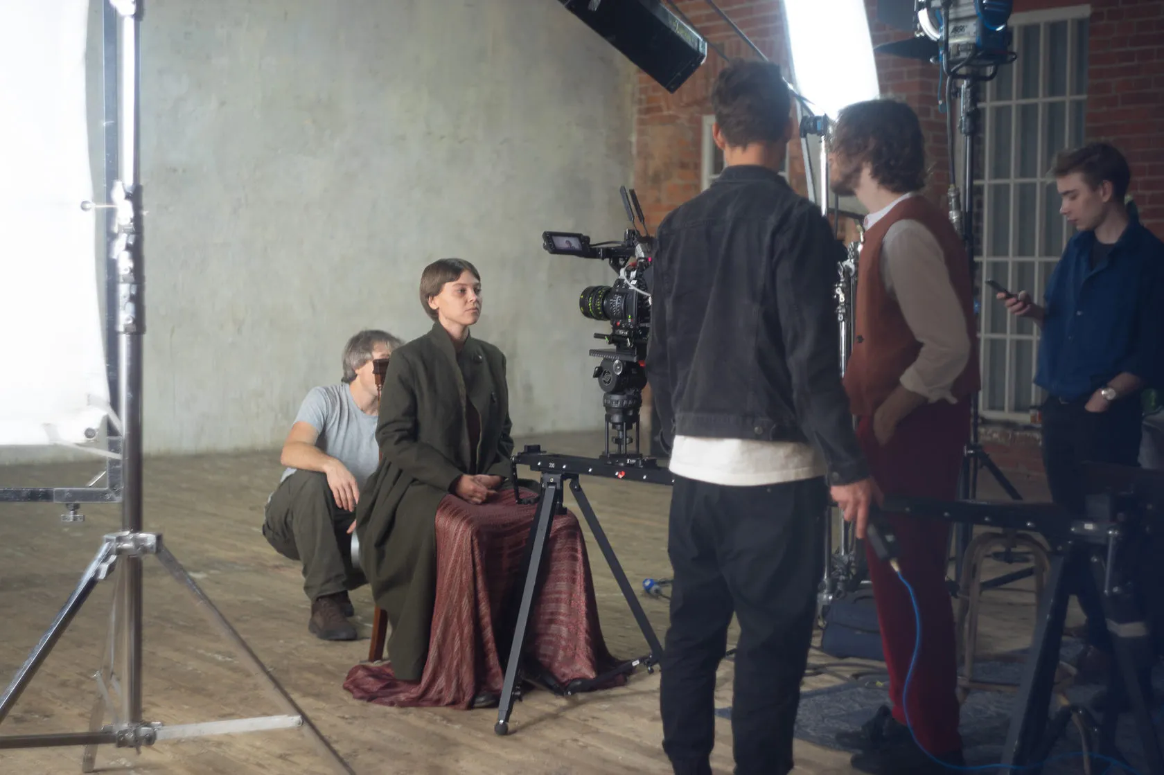 Behind the scenes: the crew sets up the camera while an actress in a long coat sits on a draped stool.