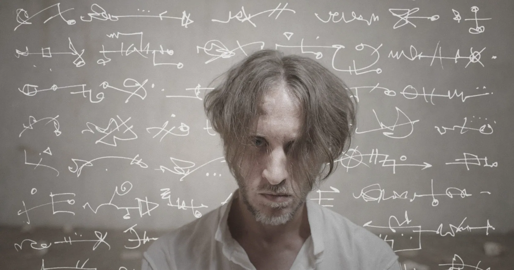 A dishevelled man in a white shirt glares in front of a wall covered in white scribbled symbols.