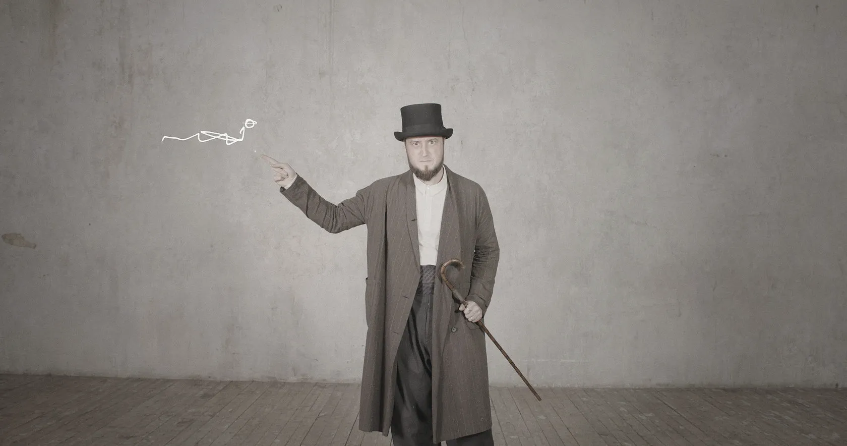 A man in a top hat and long coat leans on a cane and gestures towards a drawn squiggle.