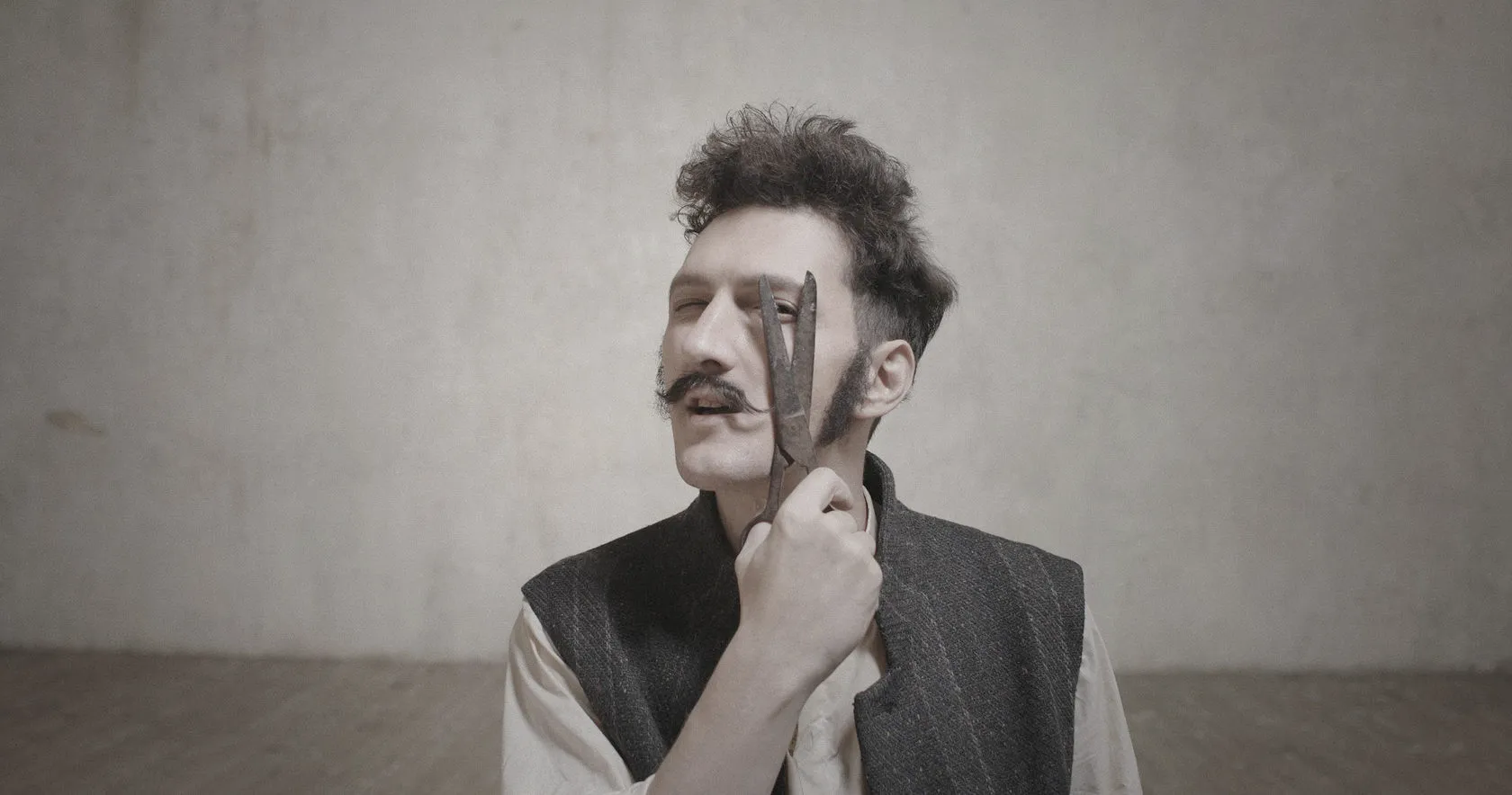 A man with a curled moustache holds an old blade to his cheek.