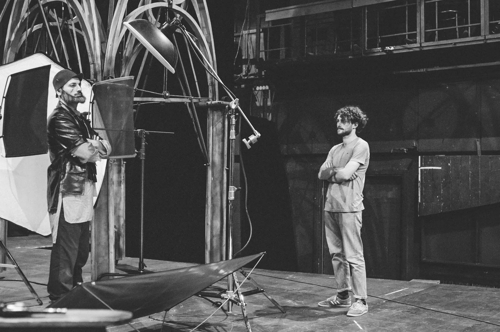 Behind the scenes: an actor in a leather coat and knitted cap waits by the light as a man looks on.