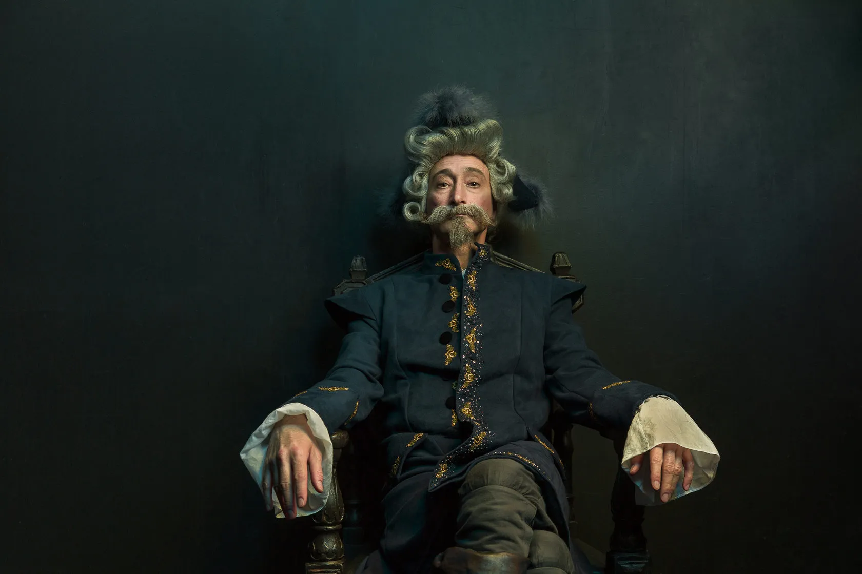 The Baron, in a fur hat and powdered wig, sits back in a carved wooden chair.