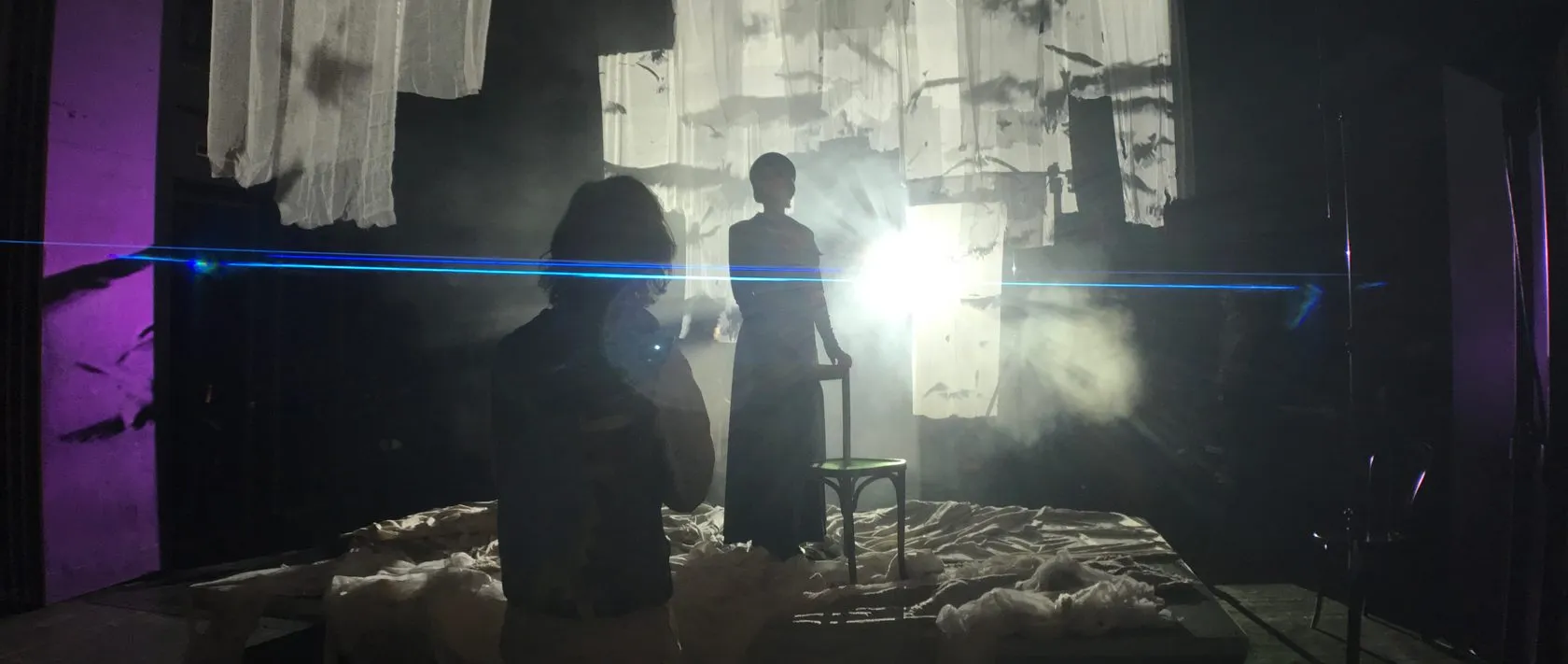 On set, a silhouetted photographer faces an actress by a chair, against backlit gauze and a laser beam.